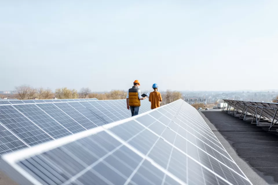 Rows of solar panels on a large rooftop with two workers in hard hats walking between rows.